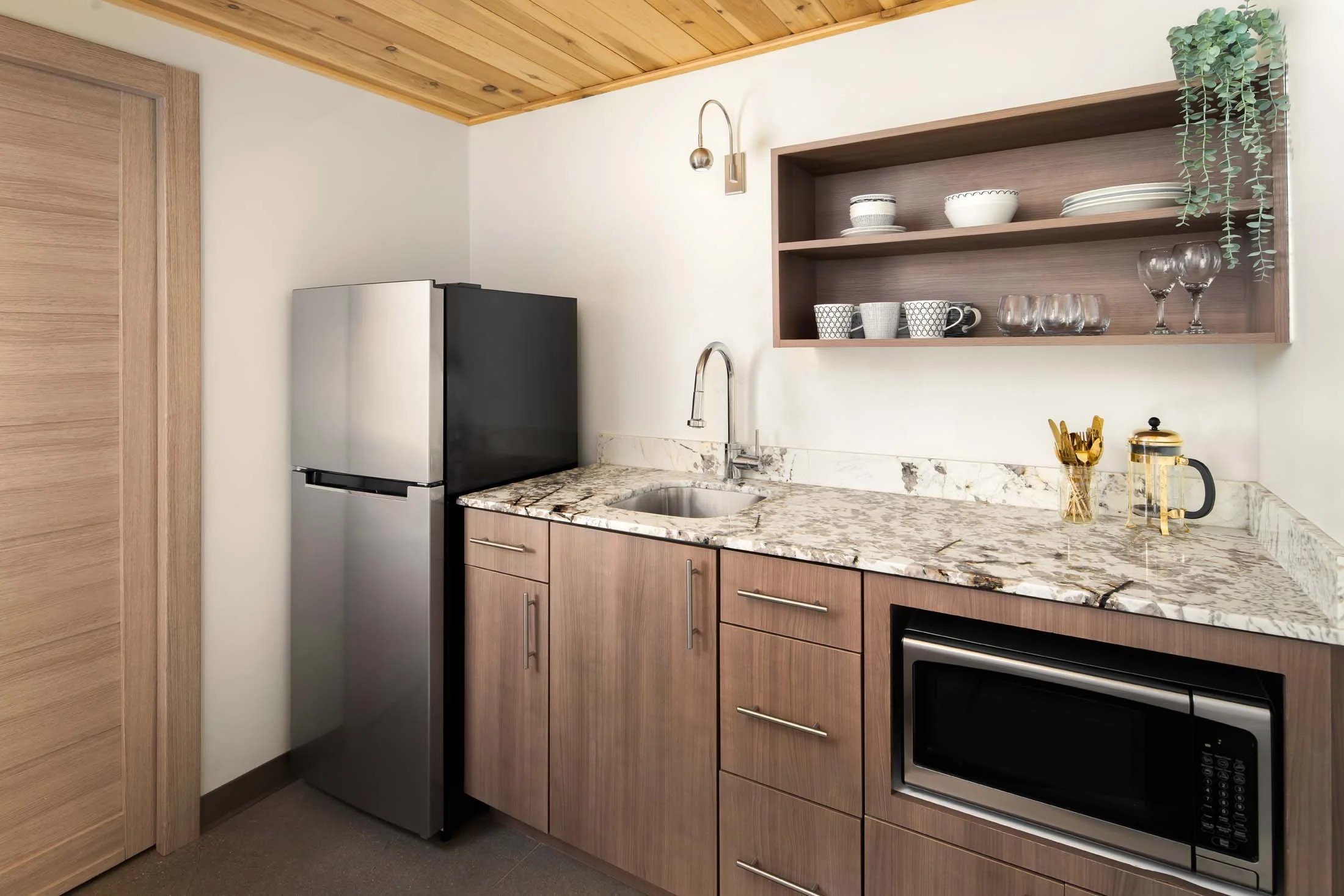 Modern kitchen with wooden cabinets, marble countertop, stainless steel refrigerator, microwave, and open shelf with dishes and glassware, decorated with green hanging plant.