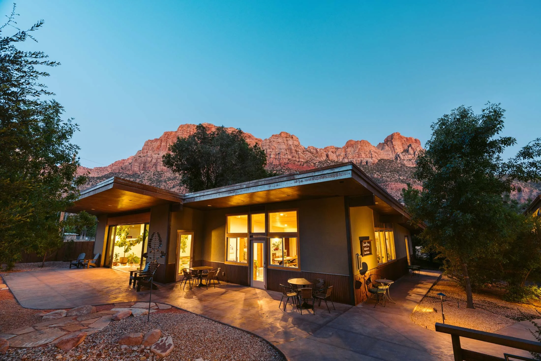 A modern house with large windows at sunset, mountains in the background, and outdoor seating area with tables and chairs.
