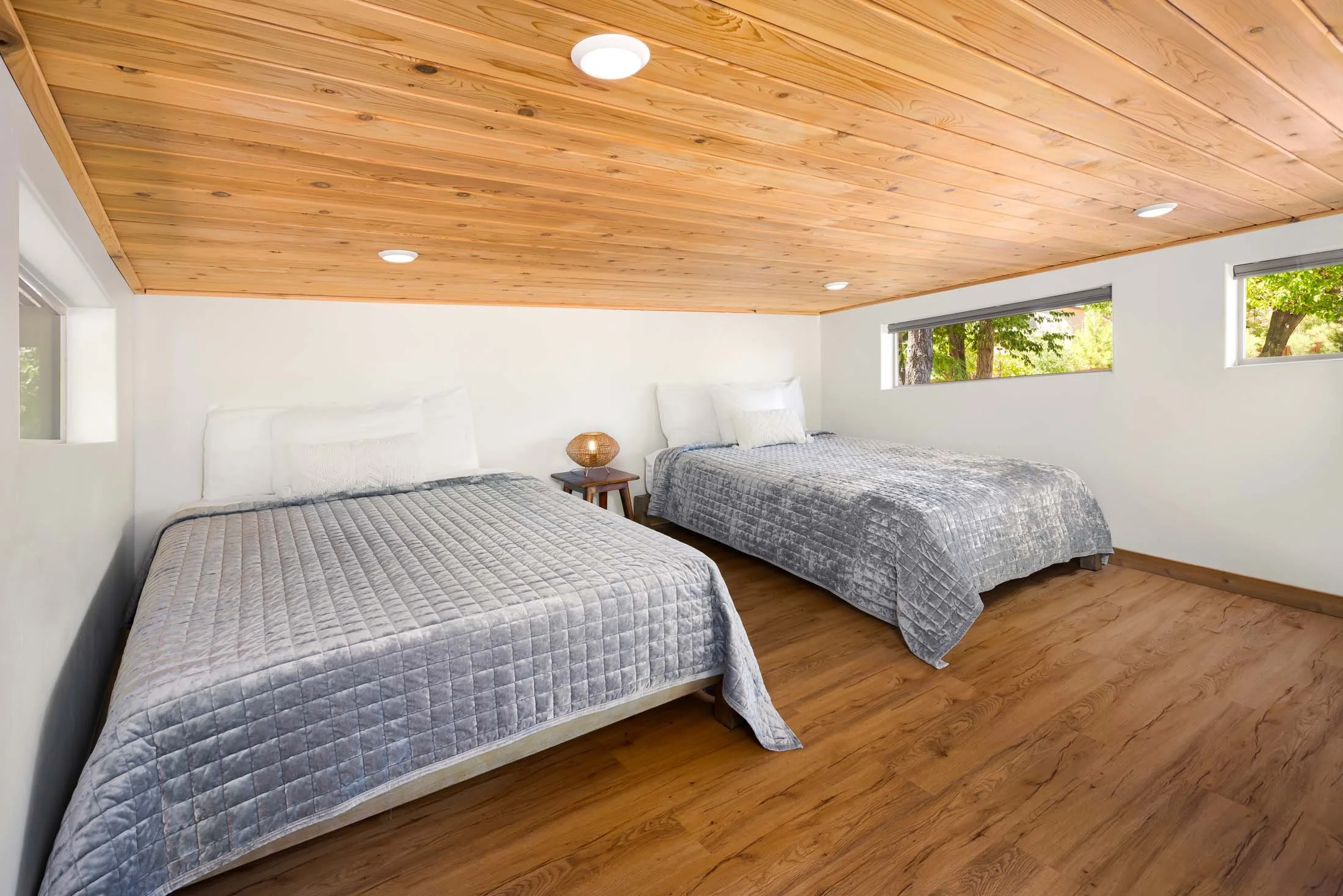 A bedroom with two beds covered in silver quilts, a wooden floor, a wooden ceiling, and three horizontal windows showing trees outside.