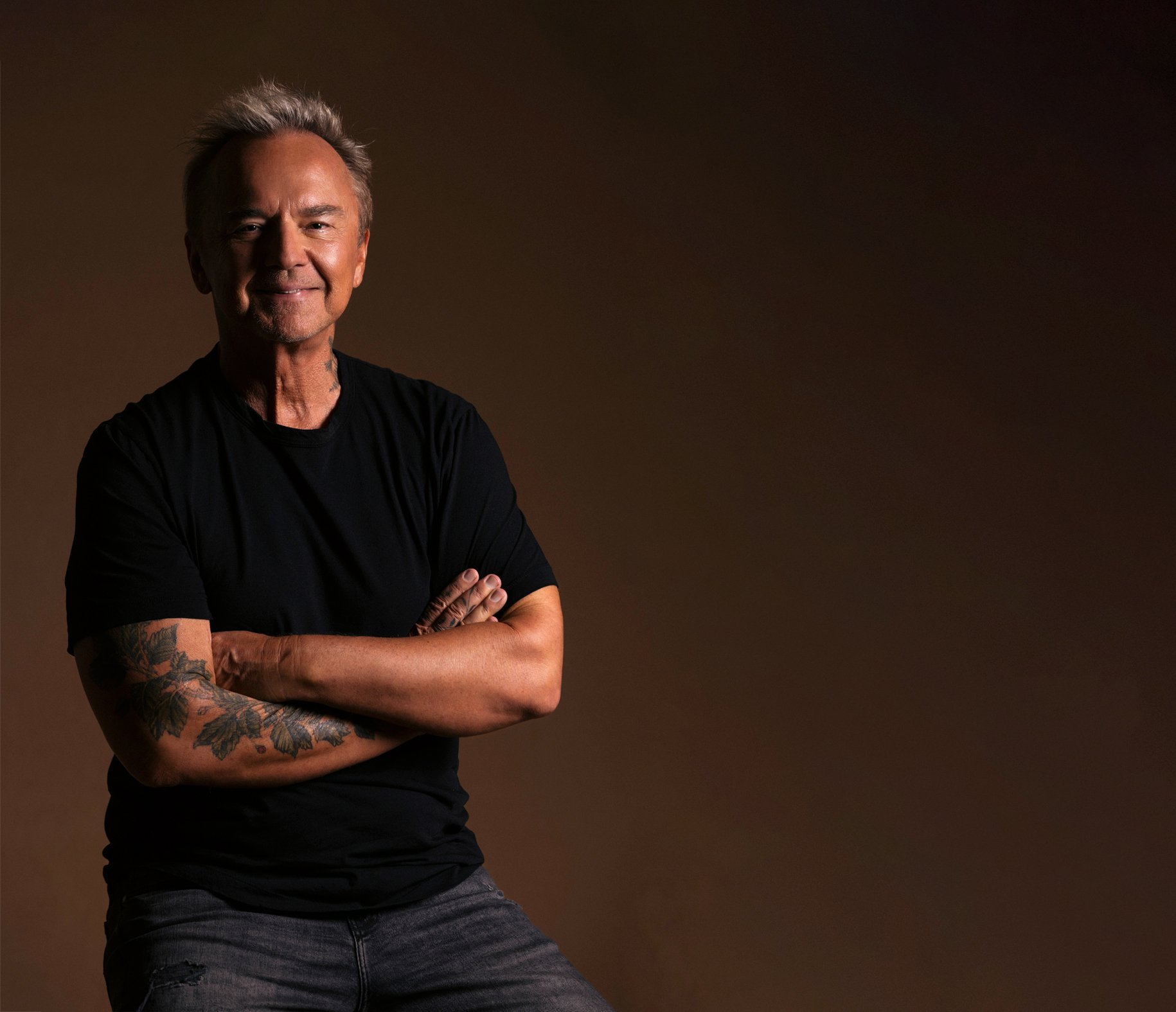 Peter Kater poses with arms crossed, wearing a black shirt against a warm brown studio backdrop.