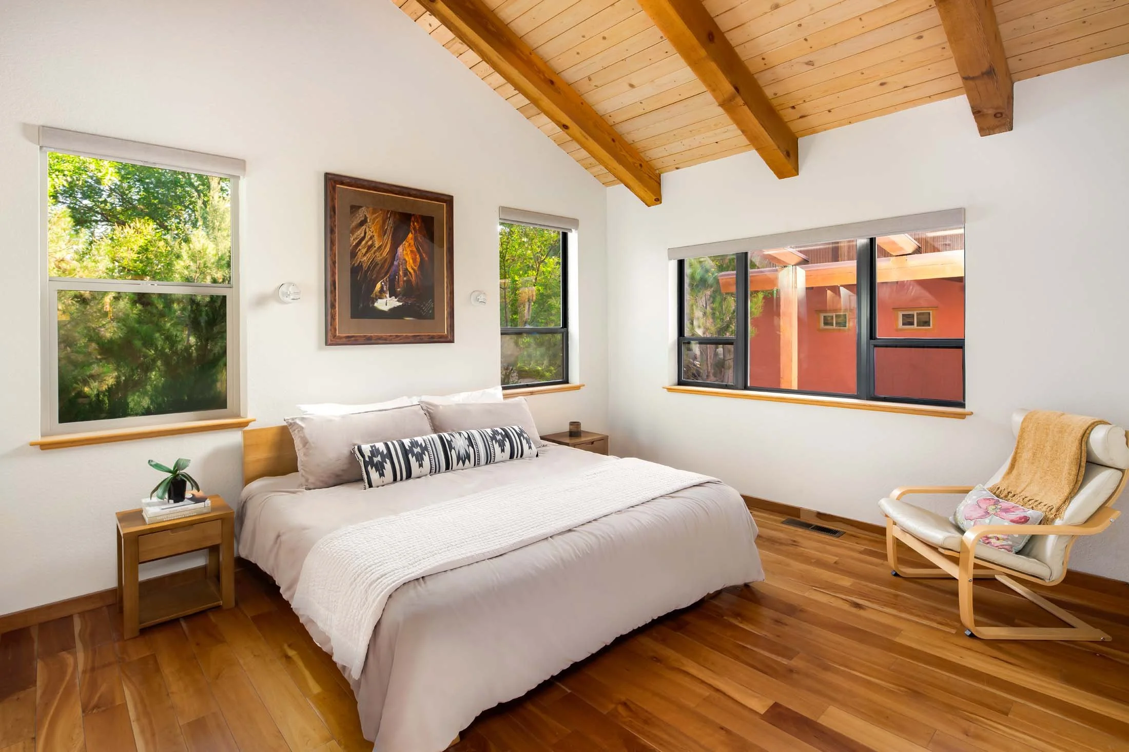A bedroom with a wooden ceiling and hardwood floor, white walls, a bed with beige bedding and pillows, a wooden nightstand with a plant, a modern armchair with a yellow blanket, and windows showing trees and a red building outside.