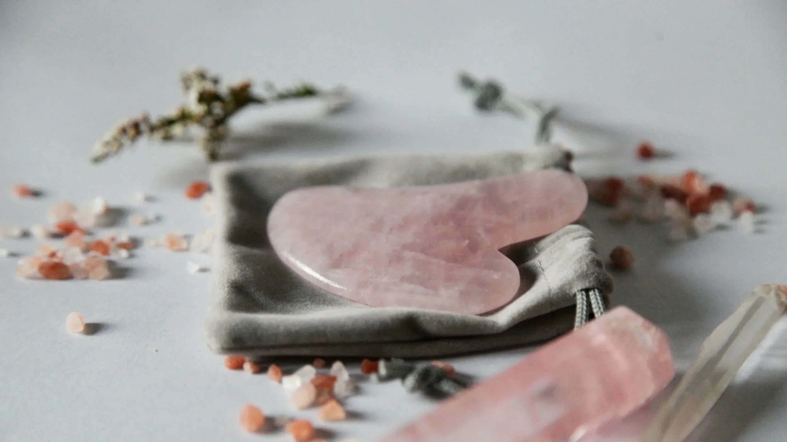 A pink heart-shaped stone on a drawstring pouch surrounded by scattered small pebbles and dried flowers on a white surface.