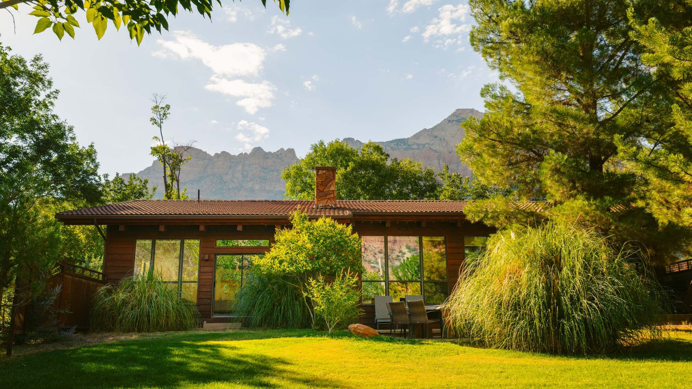 Utah wellness retreats - A wooden house with large windows is surrounded by green grass, bushes, and trees. In the background, there are mountains under a clear, partly cloudy sky.