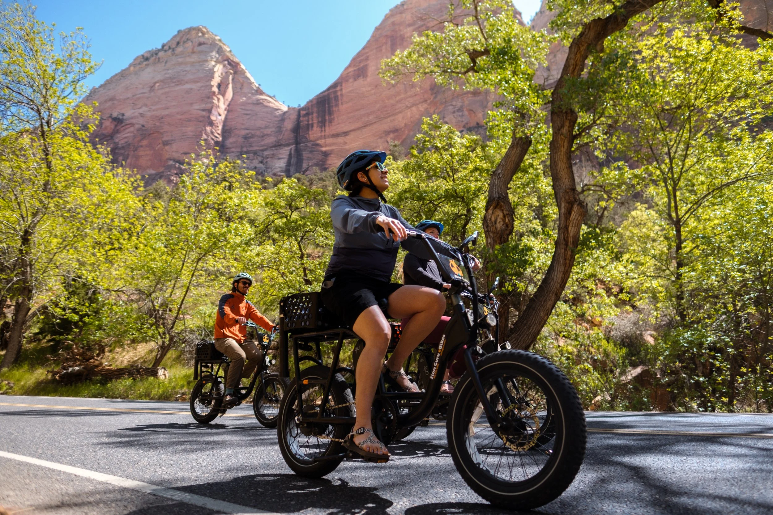Cyclists ride Zion Guru electric bikes through a tree-lined canyon beneath towering sandstone cliffs near Zion National Park, offering a scenic way to experience the greater Zion area.