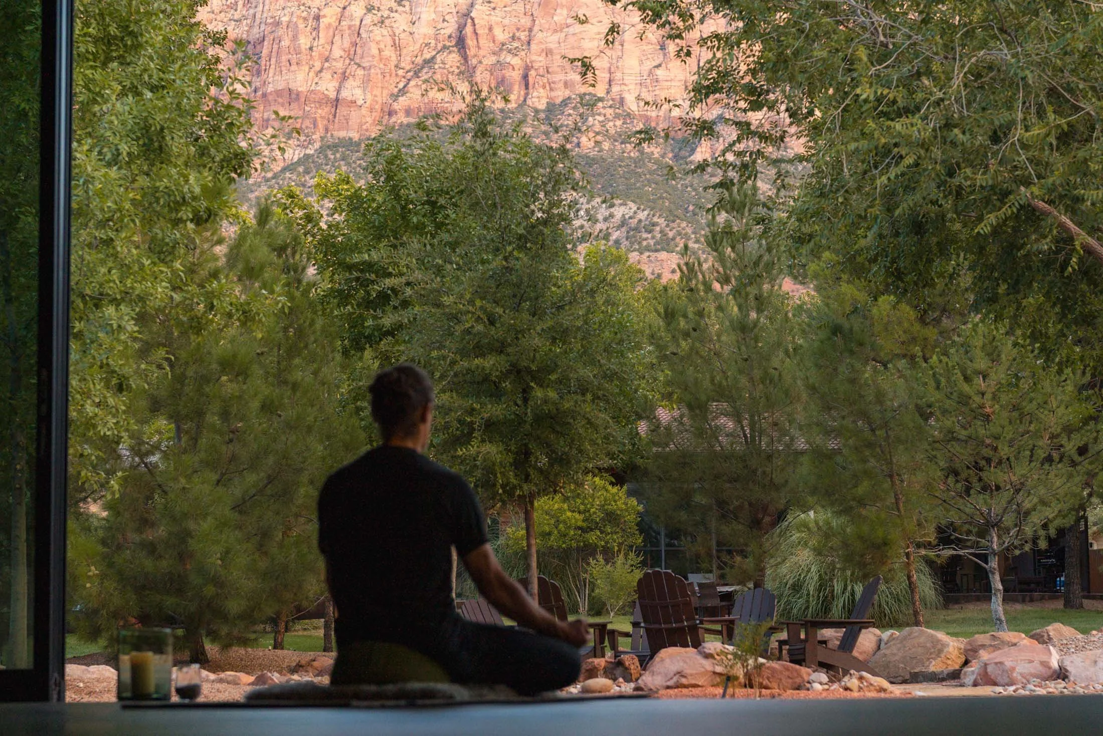 Person practicing yoga outdoors at sunset, seated in a meditative pose. The scene overlooks a lush green landscape with trees and Adirondack chairs, and a mountain range in the background.