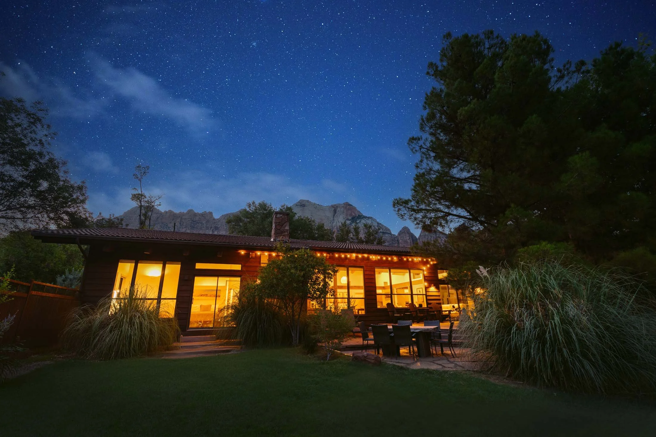 A house with large glass windows and a patio with outdoor furniture, illuminated with warm interior lights and string lights, surrounded by trees and plants under a starry night sky with mountains in the background.