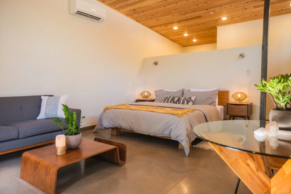 Modern bedroom with gray bed, wooden nightstands, and decorative lamps, next to a gray sofa and glass-topped table, featuring wooden ceiling and green plants.