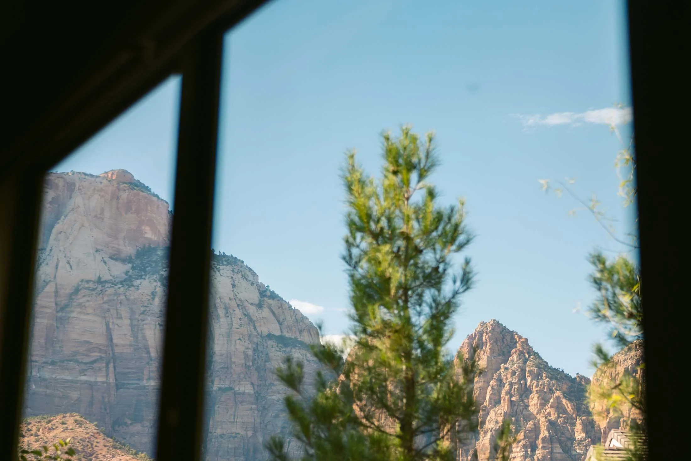 Mountain landscape viewed through a window frame with green trees in the foreground and rocky peaks in the background.