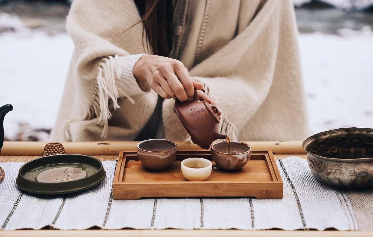 A person pouring tea from a teapot into ceramic cups on a wooden tray, with additional cups and dishes on the table outside in a snowy setting.