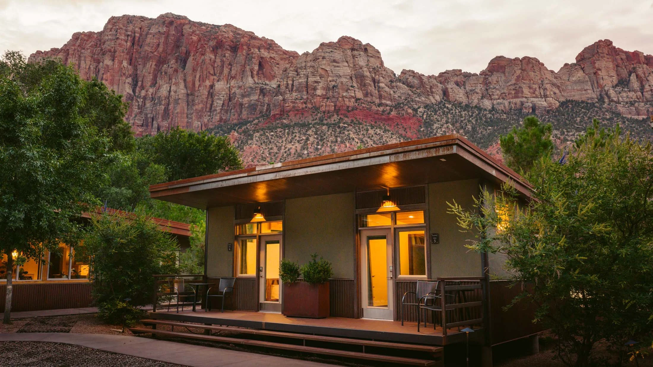 Small modern house with a wooden porch, trees, and mountains in the background at sunset.