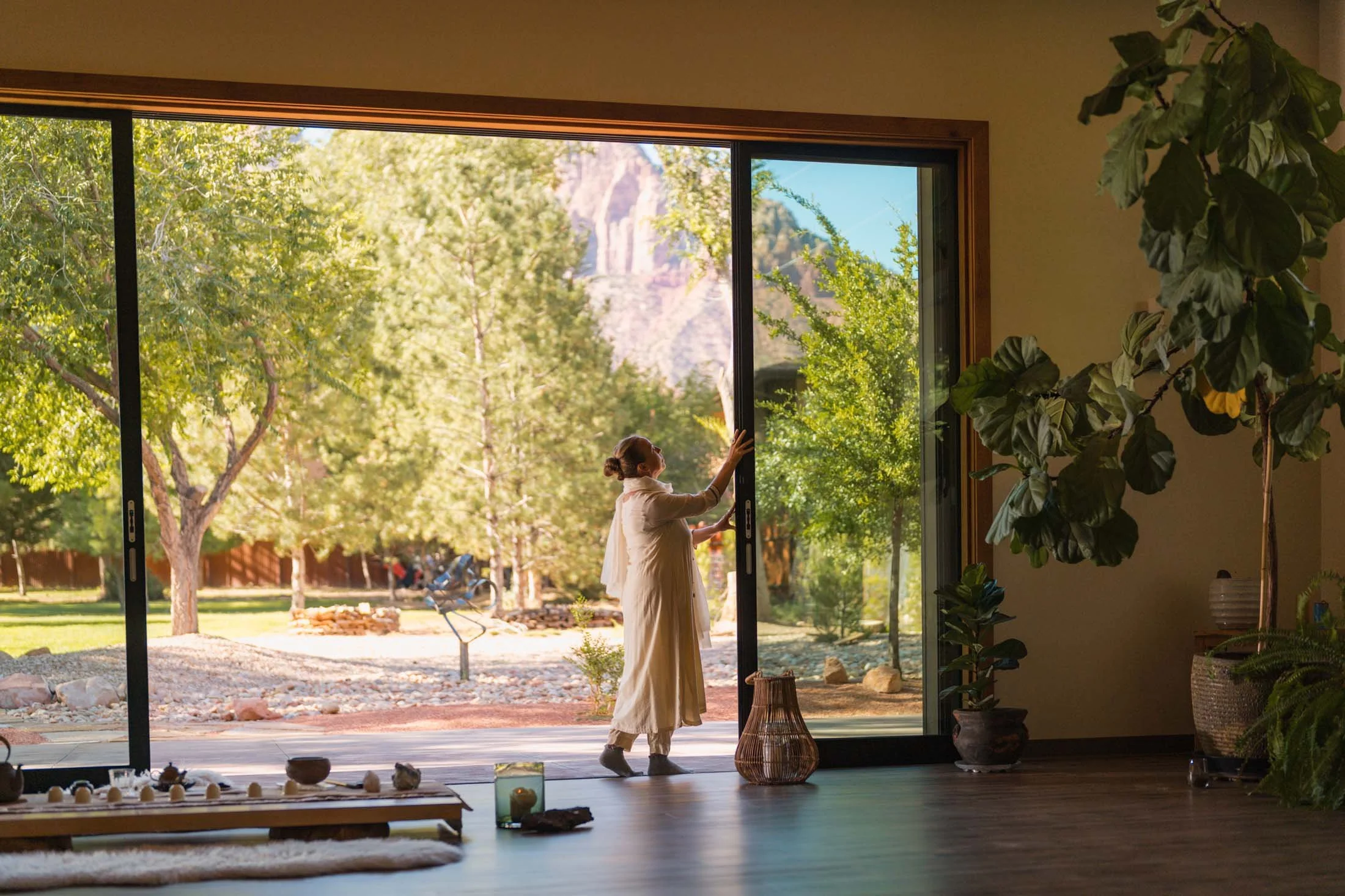 A woman opening a sliding glass door to a lush, green outdoor garden with trees and mountain in the background.