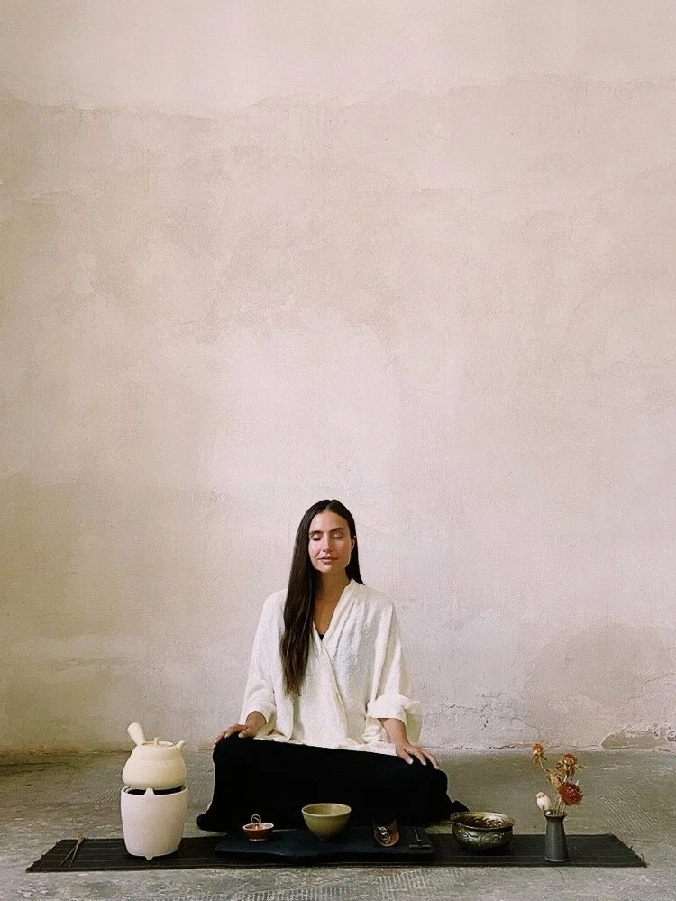 A woman sitting cross-legged on a black mat in a minimalist room with a plain white wall, surrounded by traditional Japanese tea ceremony items, including bowls, a teapot, and a flower arrangement.