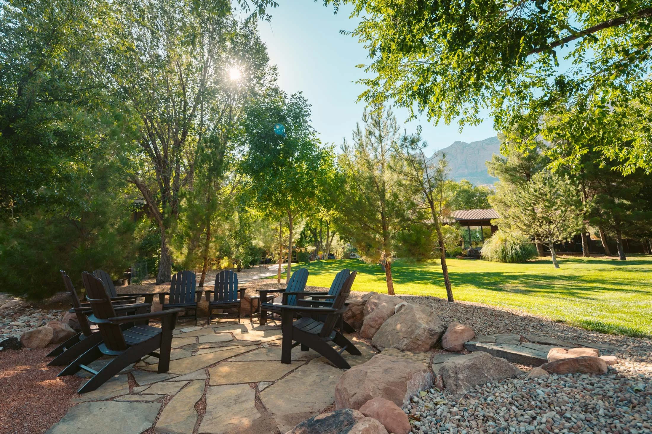 Sun shining through trees in a park with a stone patio and black Adirondack chairs