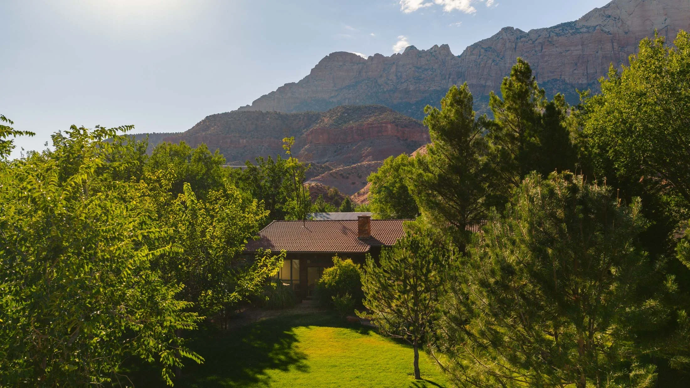 A house with a red roof is surrounded by green trees and grass, with mountains in the background under a clear sky.