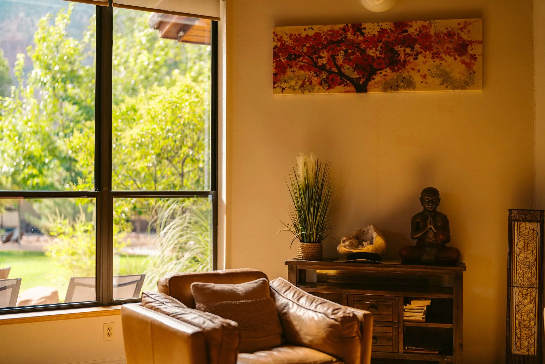 A cozy living room with sunlight streaming through a large window, a brown leather couch with pillows, a sideboard with a potted plant, a sculpture, and decorative items, a colorful tree painting on the wall, and outdoor greenery visible through the 