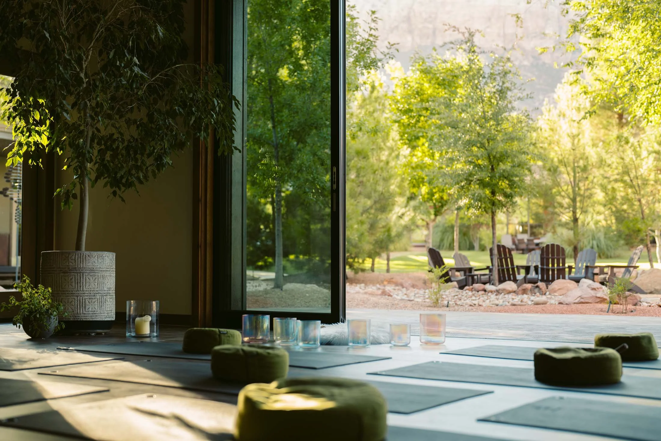 Indoor yoga or meditation space with mats, cushions, and candles, overlooking a lush outdoor garden with chairs and trees.