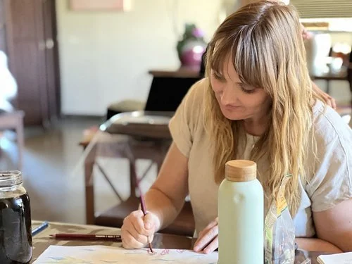 Woman with blond hair sitting at a table drawing or writing with a pencil, surrounded by art supplies and water bottles.
