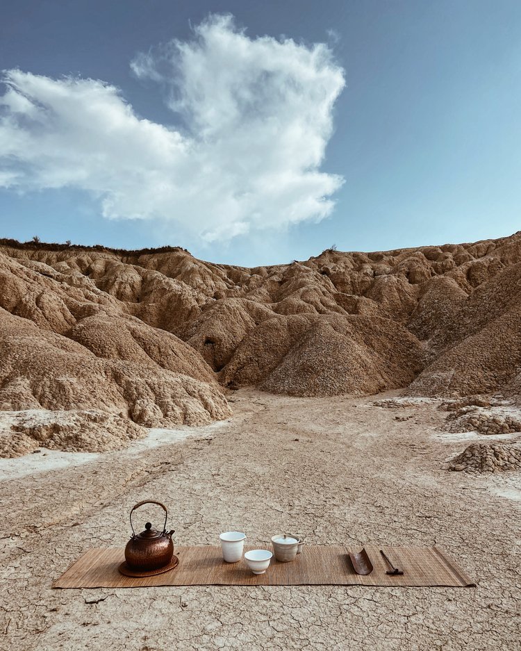 Tea set with a teapot, three cups, and utensils on a mat in a dry, rocky desert with rugged p hills and a blue sky with clouds.