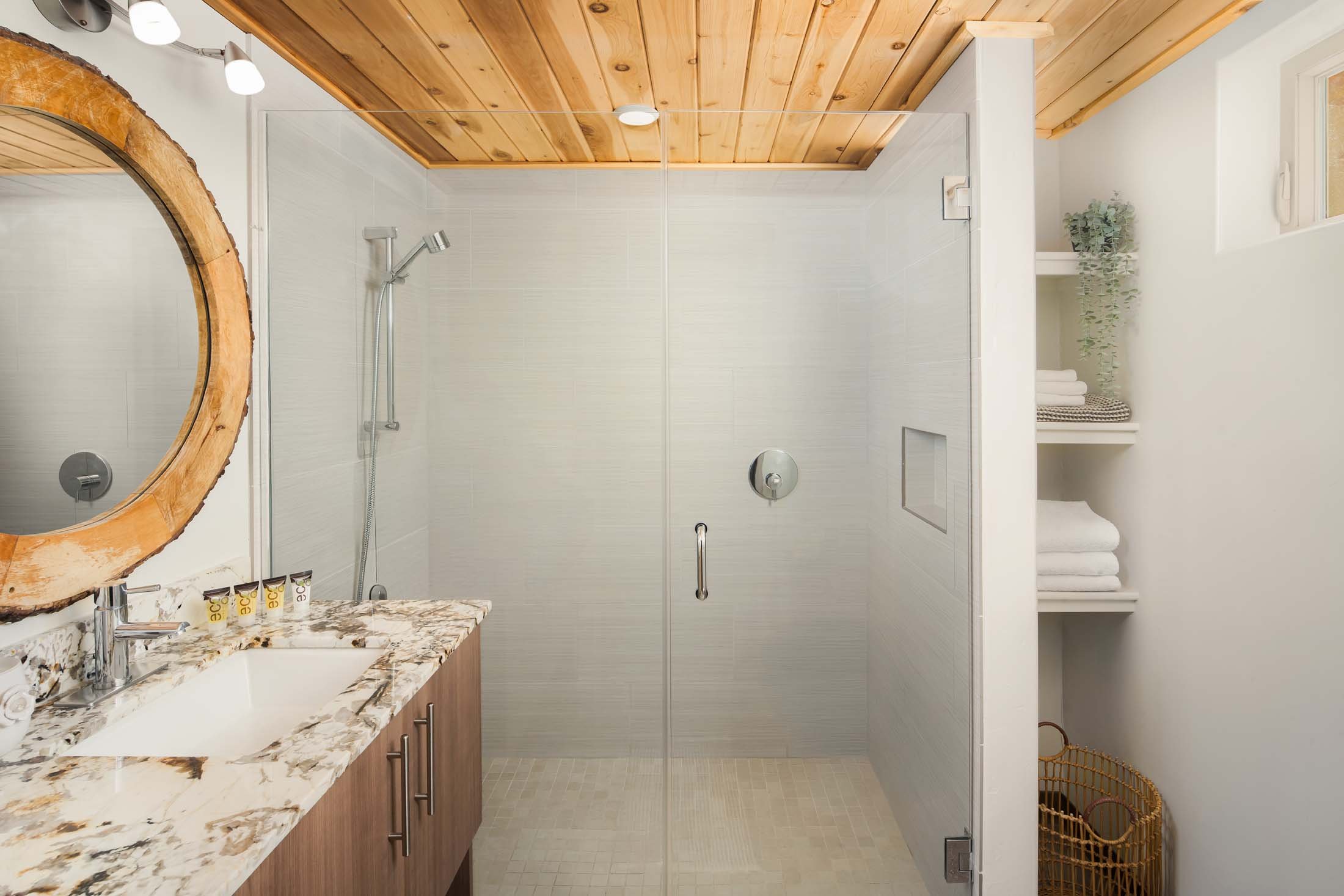 Bathroom with wooden ceiling, white tiled shower area with glass door, brown granite countertop with a sink, white shelf with towels, decorative plant