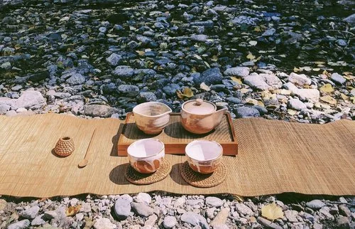 Tea cups and bowls on a tray, set on a woven mat beside a rocky river.