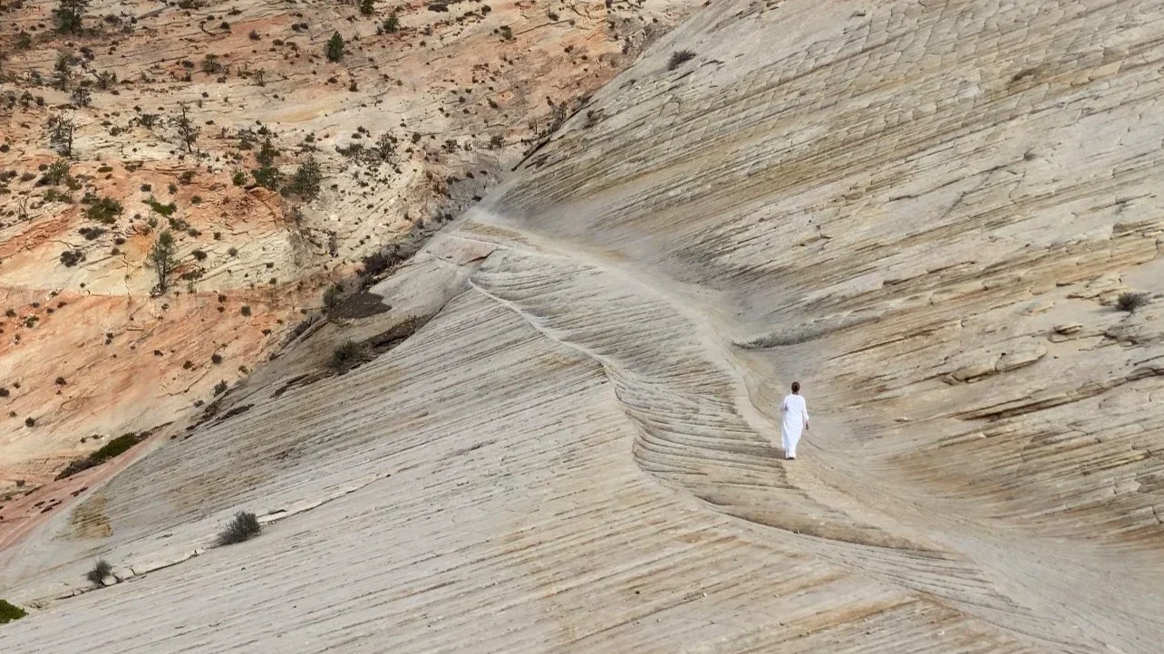A person dressed in white walking on a smooth, layered, and curving rock formation in a desert landscape.