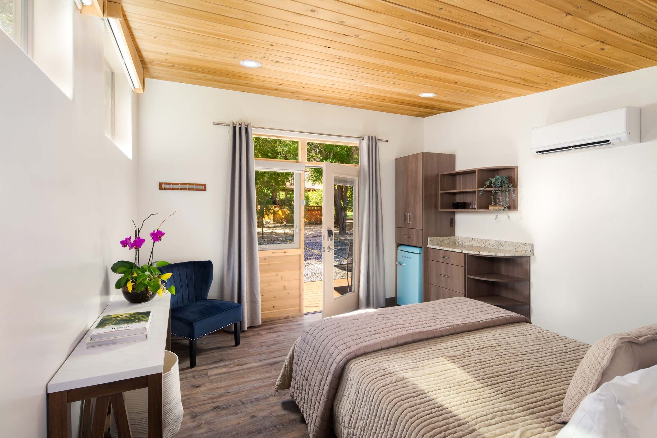 A cozy bedroom with a wooden ceiling, white walls, and a window with light gray curtains. There is a bed with beige bedding, a dark blue chair near a white desk with a potted orchid, and a small kitchenette with wooden cabinets, a mini fridge, and an air conditioning unit.