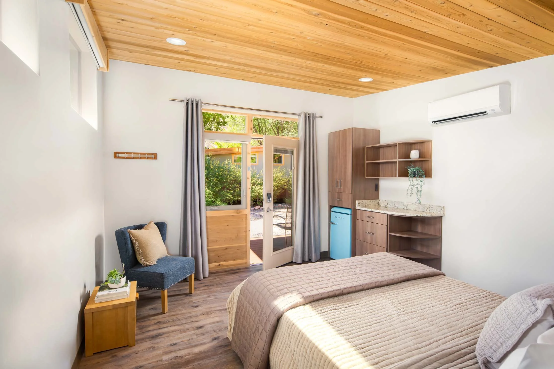 A cozy bedroom with a wooden ceiling, a bed with beige bedding, a small blue armchair with a beige pillow, a wooden side table with a plant, a door leading outside to a garden, a mini fridge, wooden cabinets, and shelves, with a white wall-mounted air conditioning unit.
