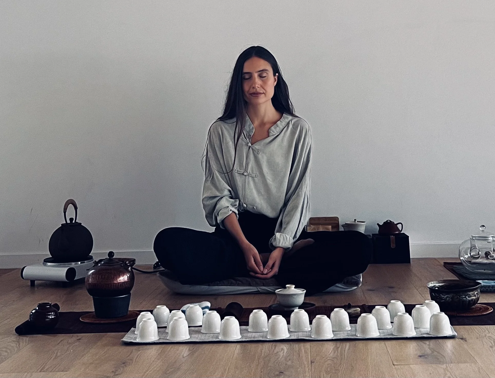 A woman practicing tea ceremony in a minimalist room, sitting cross-legged on a mat surrounded by teaware, including cups, a teapot, and other utensils.