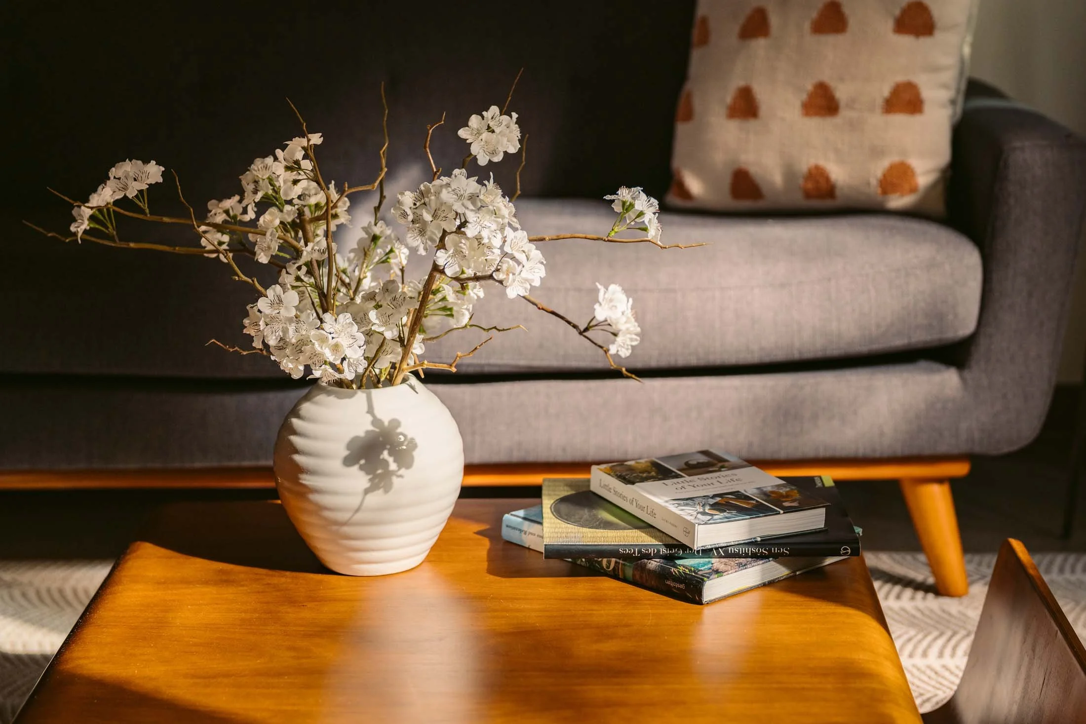 A white ribbed ceramic vase with white flowers and branches on a wooden coffee table in a living room, with a gray sofa and a patterned cushion in the background, under warm lighting.