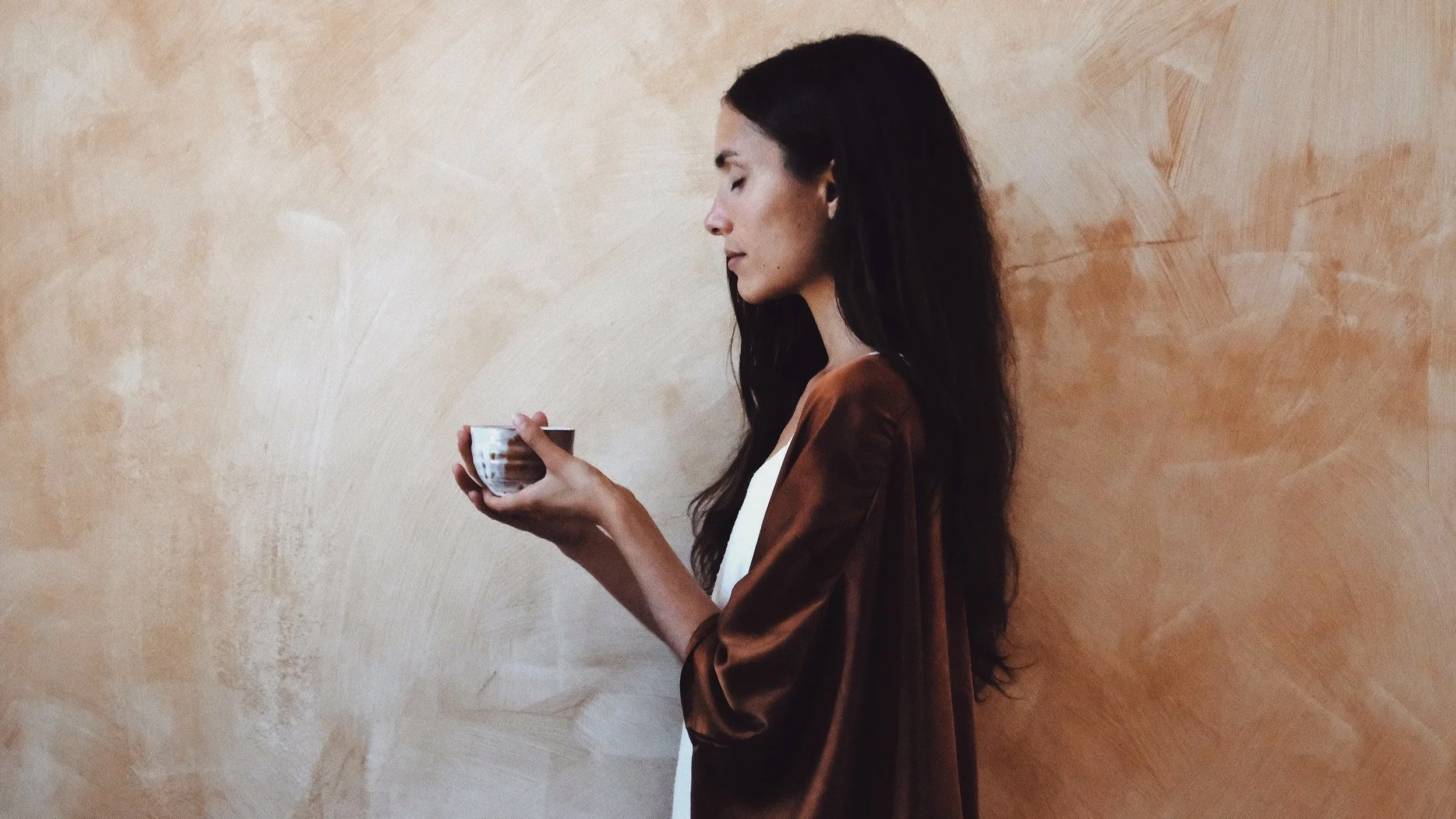 A woman with long dark hair is standing in profile against a light brown textured wall, holding a small bowl with both hands and looking at it.