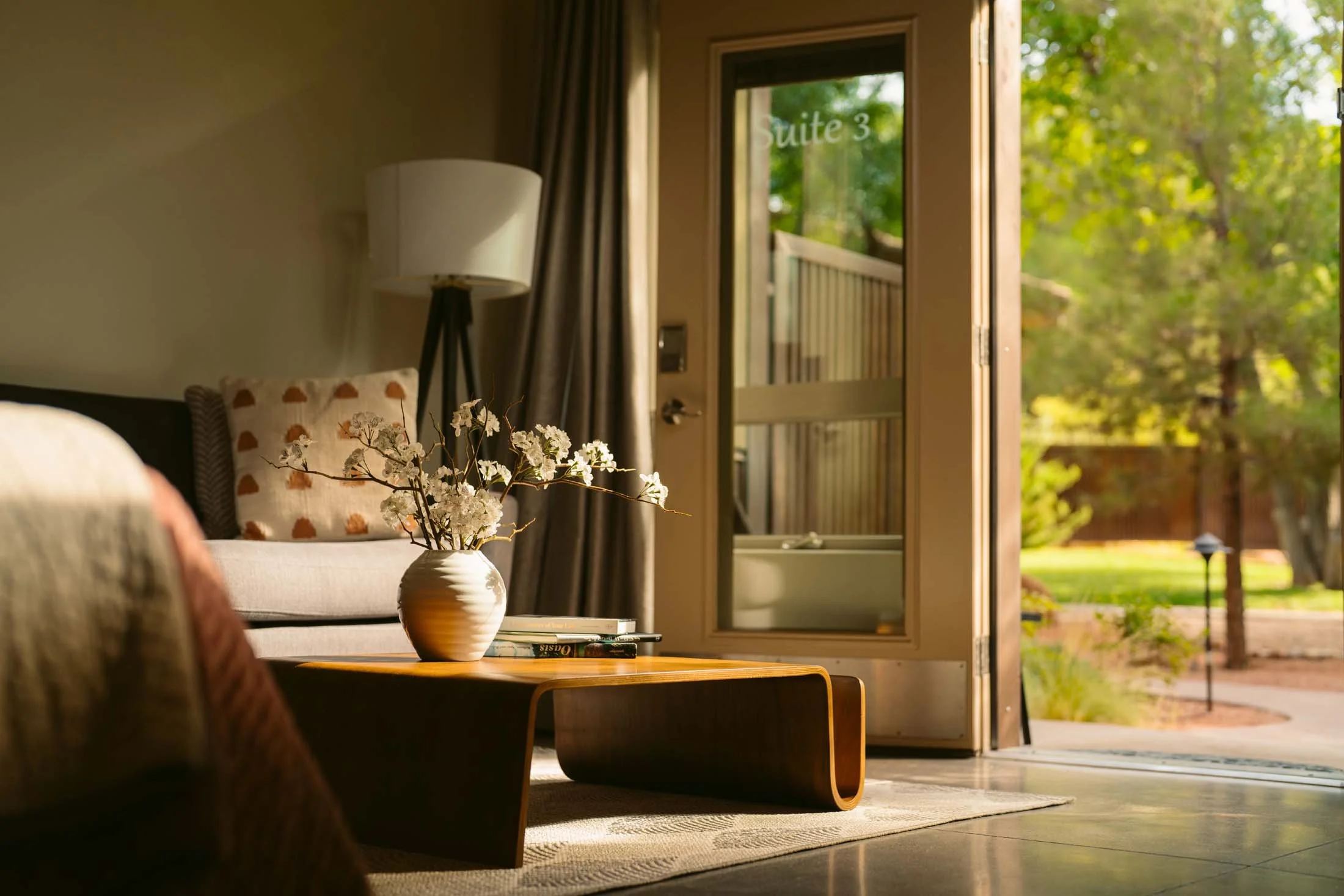 Suites Zion National Park - Living room with a coffee table, vase with white flowers, couch with decorative pillows, floor lamp, and door labeled 'Suite 3' leading outside to a garden.