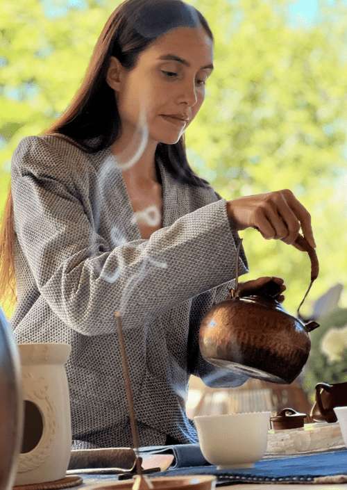 A woman in a gray blazer pours tea from a brown teapot into a white cup outdoors on a sunny day.