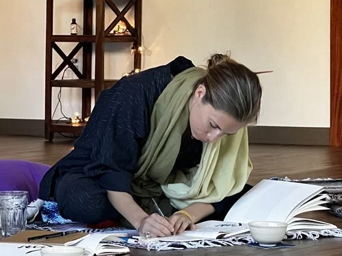 Woman sitting on the floor, writing in a notebook with open magazines or books around her.