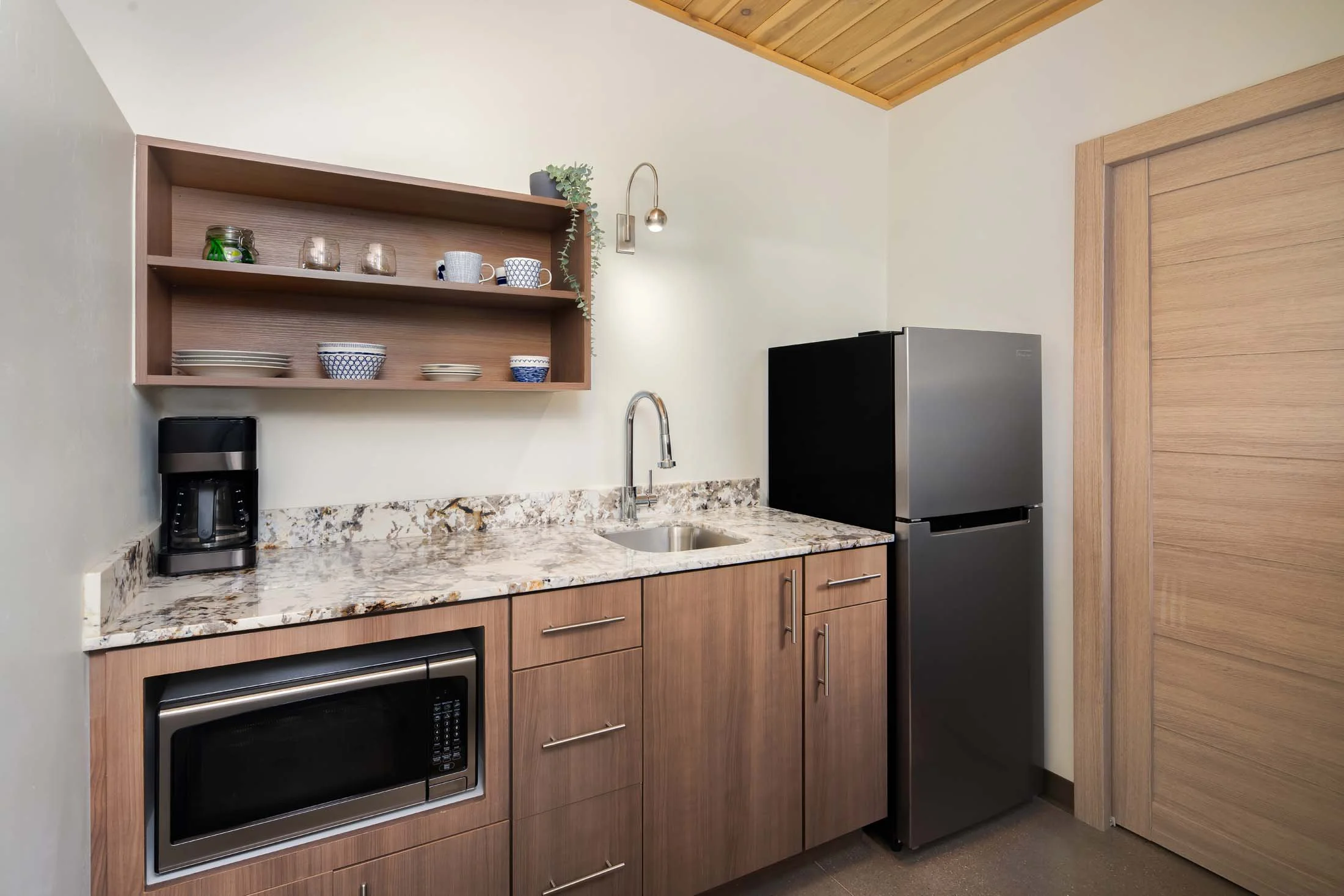 Small kitchen area with wooden cabinets, granite countertop, a mini refrigerator, microwave, coffee maker, open shelves with dishes, a sink, and a wooden ceiling panel.