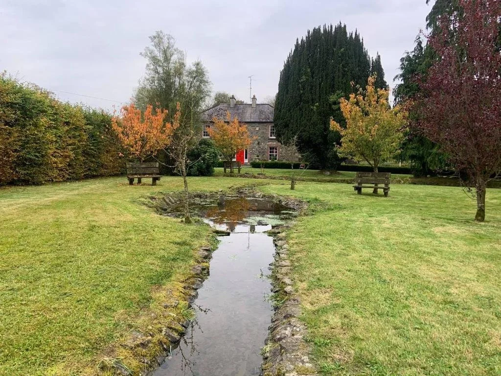 A park with a small stream running through the grass, surrounded by trees with autumn leaves, benches, and a large house in the background under an overcast sky.