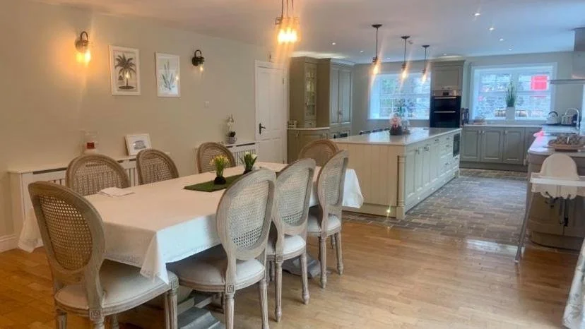 A spacious kitchen and dining area with a long dining table, eight beige chairs, a white kitchen island, cabinetry, and large windows with a view outside.