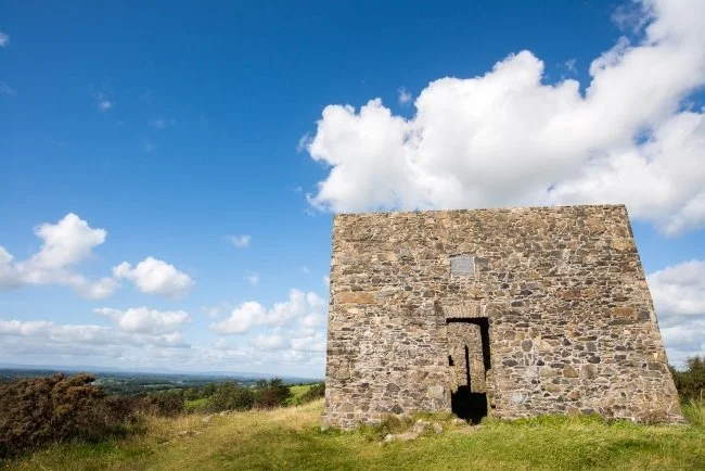 A large stone structure with a rectangular shape and a small opening at the bottom, situated on grassy land under a partly cloudy sky.