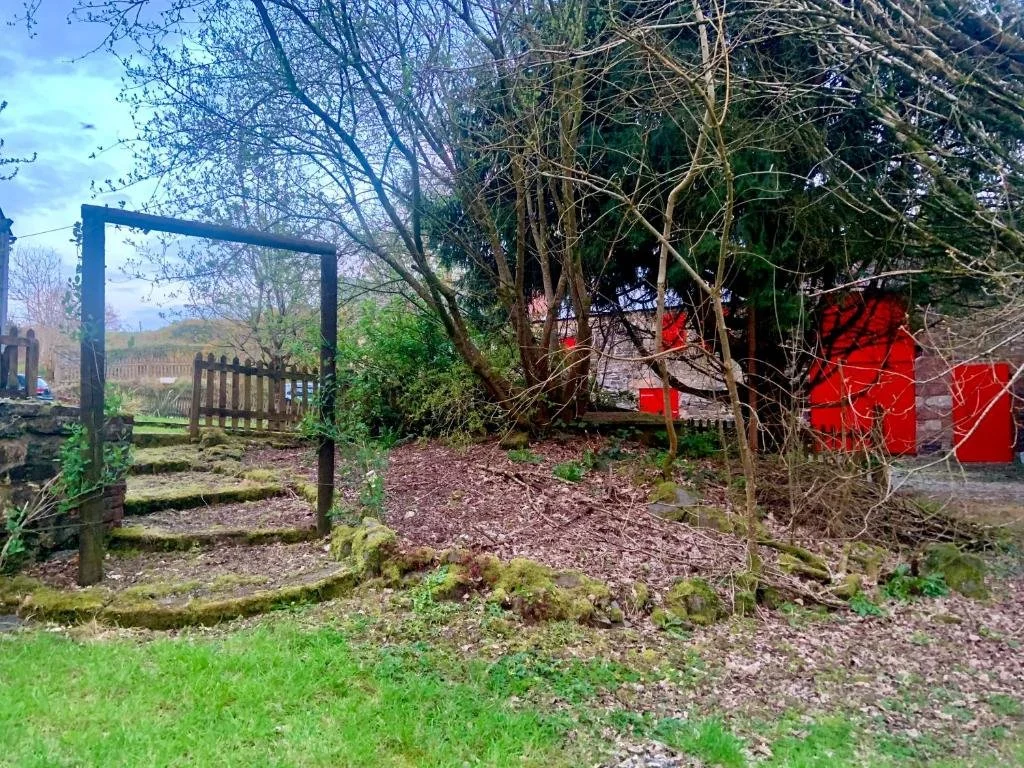A garden scene with a stone stairway leading upwards, a wooden gate on the left, and a densely wooded area with a tree and leafless branches on the right. In the background, there are red objects and a white wall.