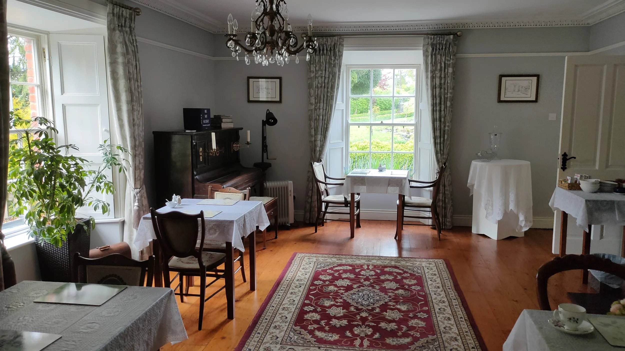 A vintage-style dining room with white walls, large windows with patterned curtains, and wooden flooring. There are multiple tables with white tablecloths, a piano in the corner, a chandelier hanging from the ceiling, and a red patterned rug on the floor.