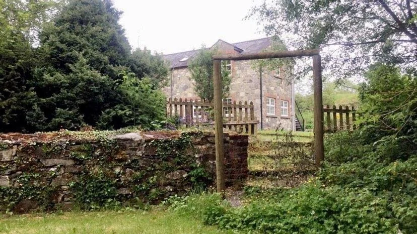 An old stone house in the background with a wooden fence and a makeshift wooden gate in the foreground, surrounded by greenery.