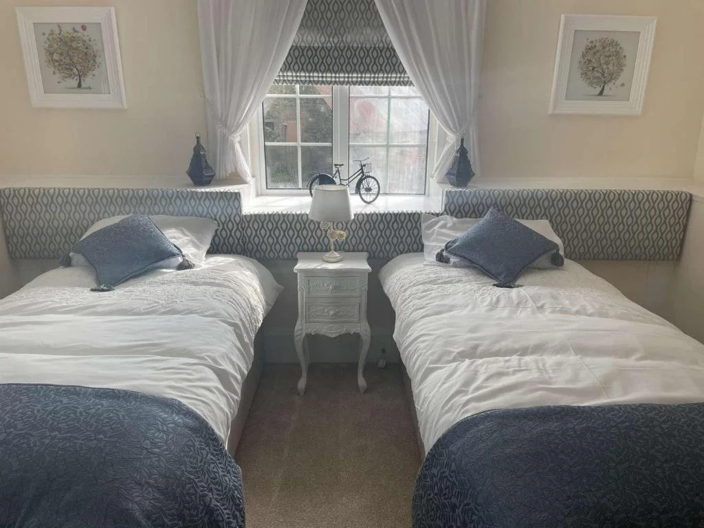 Two twin beds with white bedding and decorative pillows, separated by a small white nightstand with a lamp. A window with curtains and a valance behind the beds displays a bicycle on the windowsill, with framed artwork of trees on the wall.