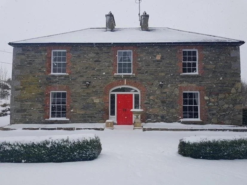 Stone house with red door, snow-covered ground, and bushes in front, during winter.