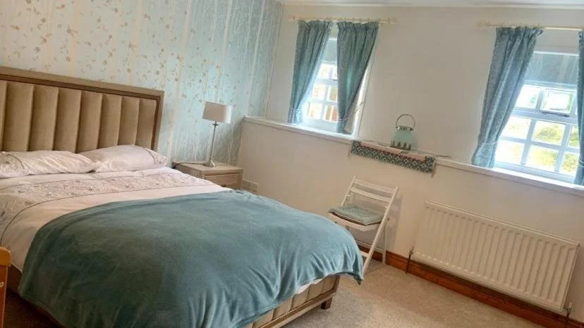A cozy bedroom with a bed, nightstand, and lamp, featuring light-colored walls and blue curtains by the windows.