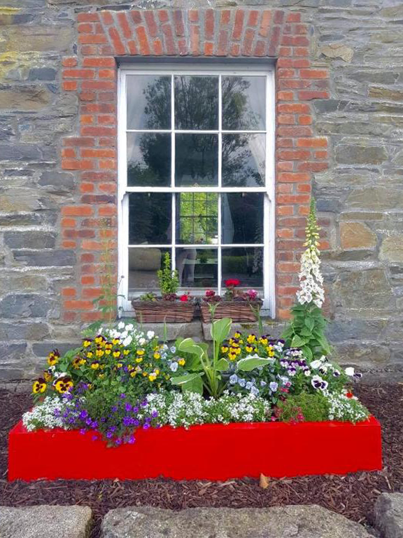 A window with red brick trim set into a stone wall. In front of the window is a vibrant red flower box filled with colorful flowers, including pansies, violas, and other small blooming plants, with some greenery and a tall white flower plant on the side.