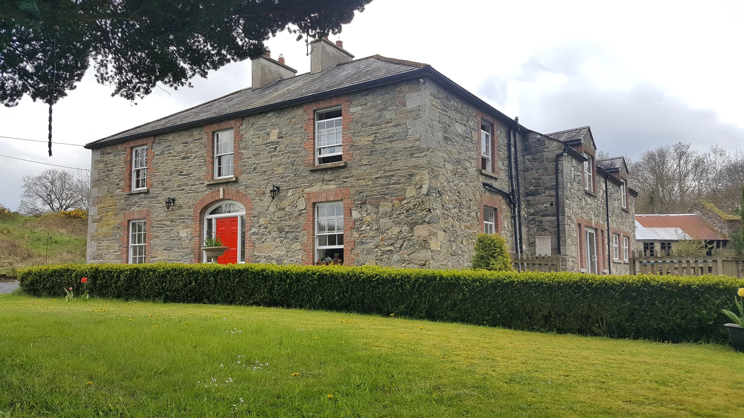 A large stone house with six windows, a red front door, and a lush green lawn with a neatly trimmed hedge in front.