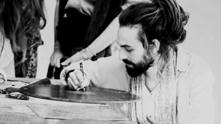 Un homme avec des cheveux longs et une barbe, assis à une table, en train de peindre ou de dessiner sur un disque en vinyle.
