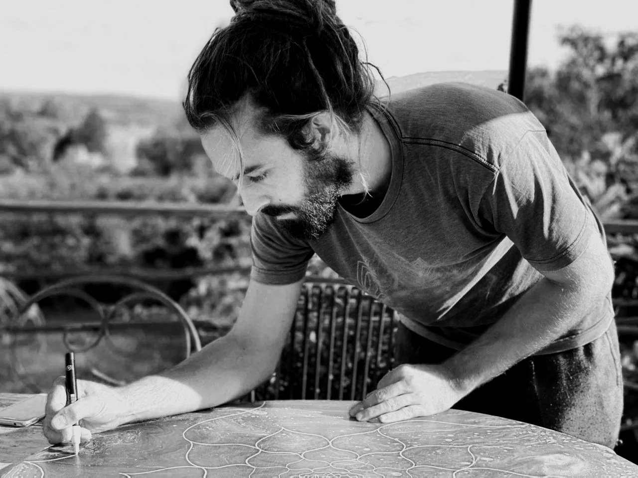 Un homme avec une barbe et des cheveux longs, portant un t-shirt, écrit ou dessine sur une table en plein air.
