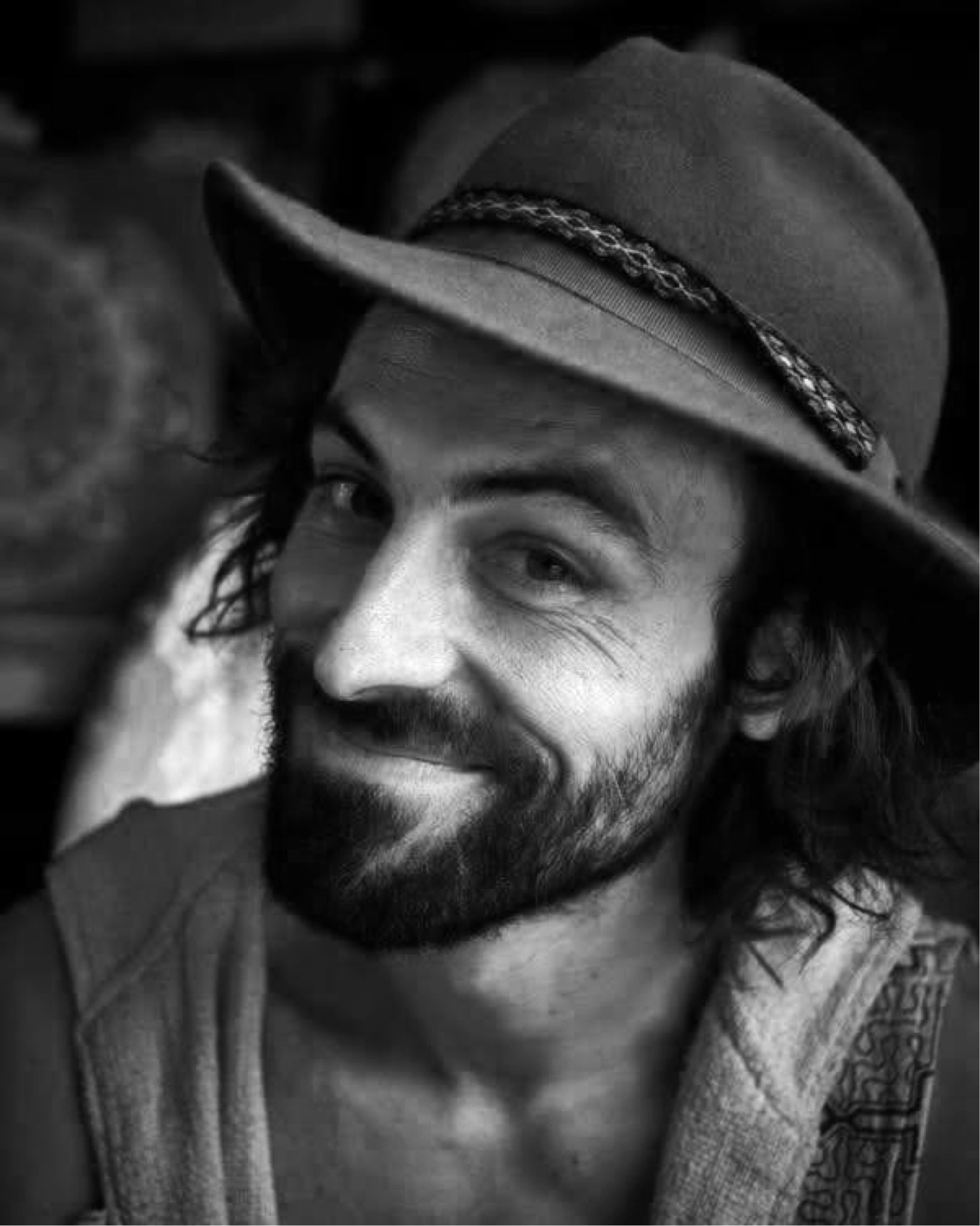 Un homme avec une barbe, portant un chapeau, souriant de côté dans un portrait en noir et blanc.