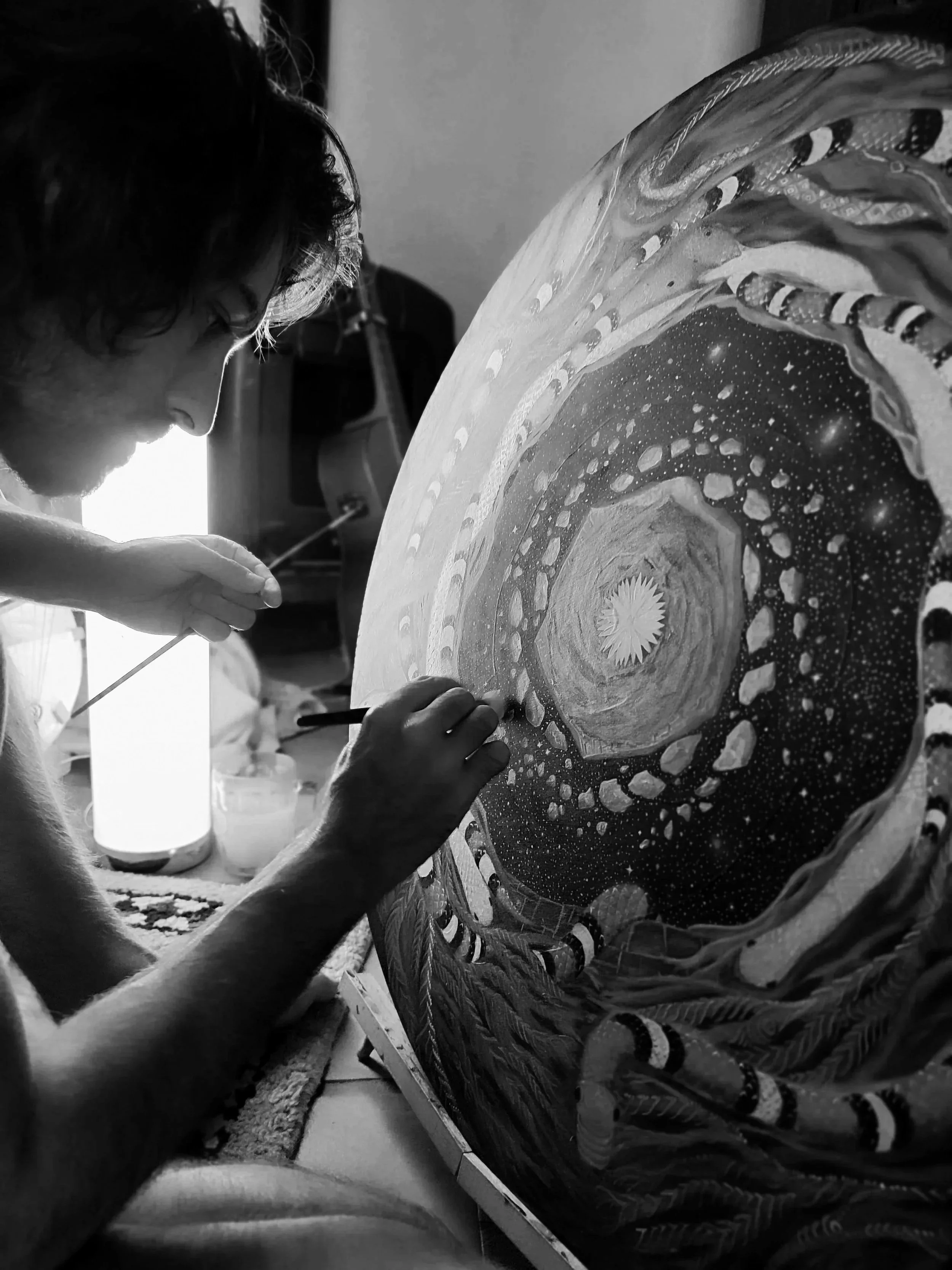 Une personne en train de peindre une toile ronde représentant une scène cosmique ou galactique, avec des détails tels que des étoiles, des planètes et un centre lumineux, dans un environnement intérieur.