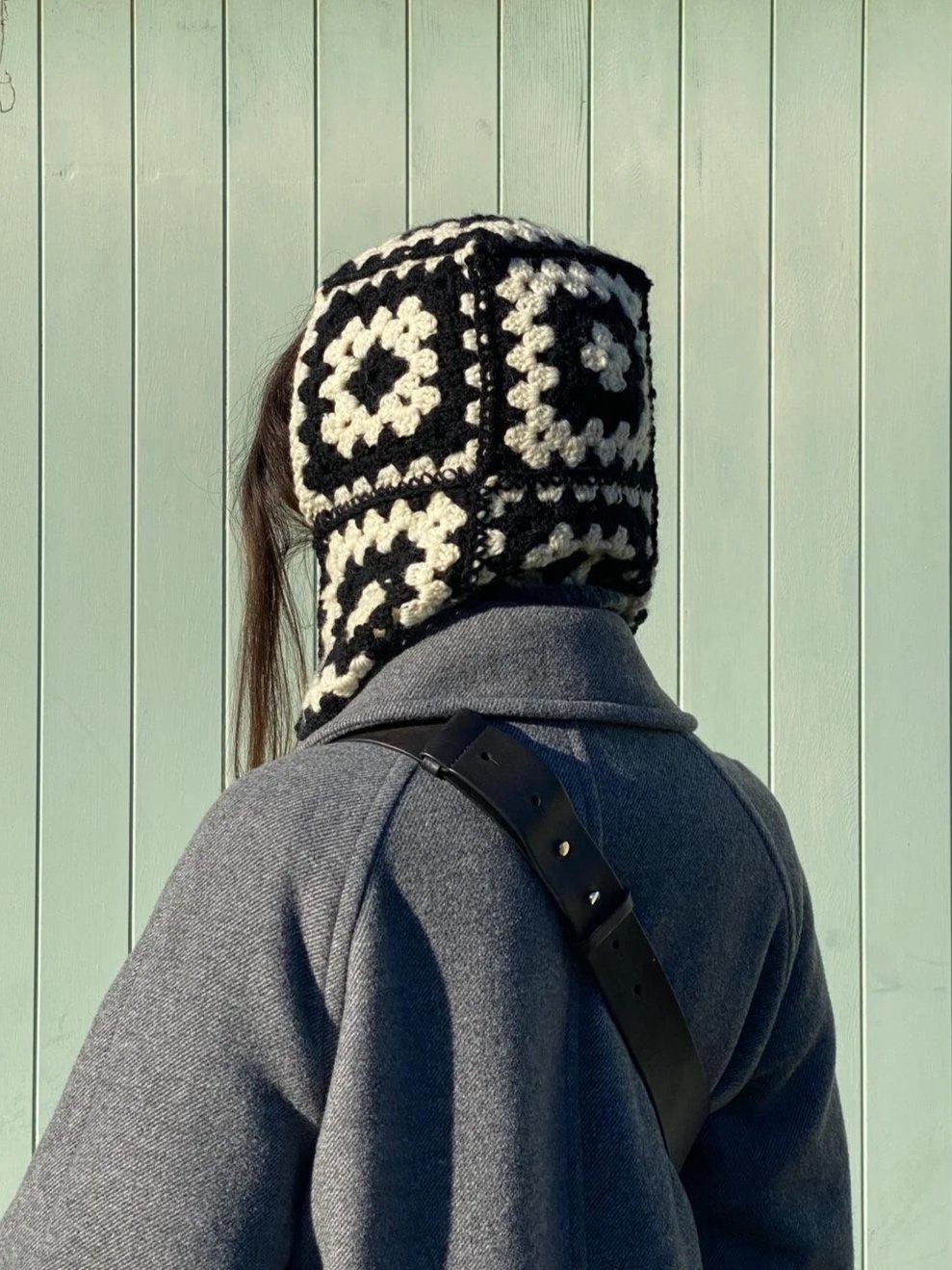 Person wearing a black and white crocheted hood, a gray coat, and a black strap over the shoulder standing in front of a pale green wooden wall.